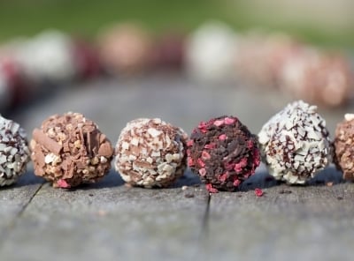 The image shows a wooden surface with a row of six round, bite-sized chocolate truffles, each coated in various toppings like chocolate, nuts, and sprinkles. The background is blurred with hints of greenery.